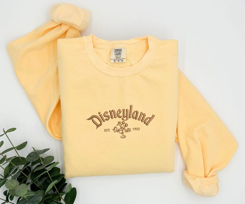 Puede incluir: Una sudadera amarilla clara con la palabra "Disneyland" bordada en escritura marr&oacute;n, junto con un gr&aacute;fico de Mickey Mouse y el a&ntilde;o "1955". La camisa de manga larga est&aacute; hecha de un material suave.