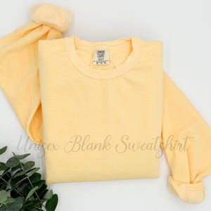 May include: A light yellow unisex blank sweatshirt with the text "Unisex Blank Sweatshirt" in cursive. The sweatshirt has a crew neck and long sleeves. The Comfort Colors tag is visible.
