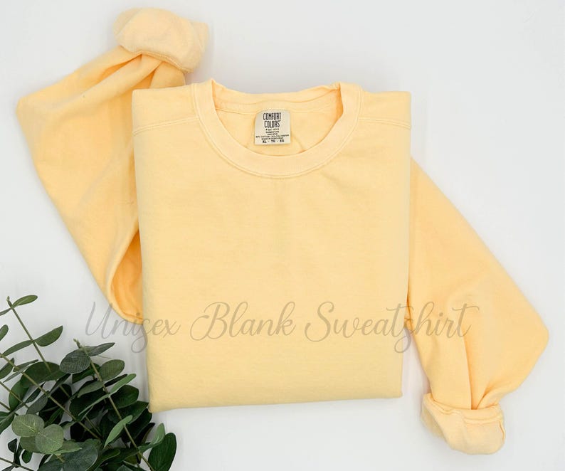 May include: A light yellow unisex blank sweatshirt with the text "Unisex Blank Sweatshirt" in cursive. The sweatshirt has a crew neck and long sleeves. The Comfort Colors tag is visible.