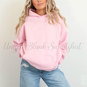 May include: A light pink unisex hoodie with a front pocket and a drawstring hood. The sweatshirt is blank and has the words "Unisex Blank Sweatshirt" in a cursive font. The model is wearing blue jeans.