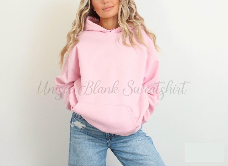 May include: A light pink unisex hoodie with a front pocket and a drawstring hood. The sweatshirt is blank and has the words "Unisex Blank Sweatshirt" in a cursive font. The model is wearing blue jeans.
