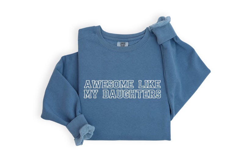 以下が含まれることがあります： 「AWESOME LIKE MY DAUGHTERS」の文字が白でプリントされた青いスウェットシャツ。長袖シャツはきれいに折りたたまれ、袖と首元が見えます。生地は柔らかく快適に見えます。