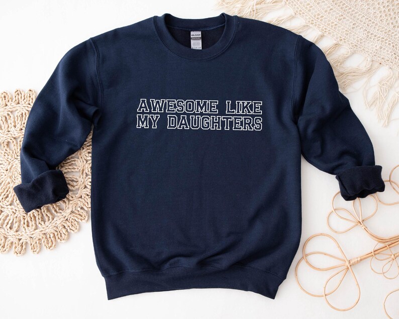 以下が含まれることがあります： 「AWESOME LIKE MY DAUGHTERS」の白文字がプリントされたネイビーブルーのスウェットシャツ。クルーネックのスウェットシャツは、リブ編みの袖口が付いた長袖です。衣類はニュートラルな背景に表示されています。