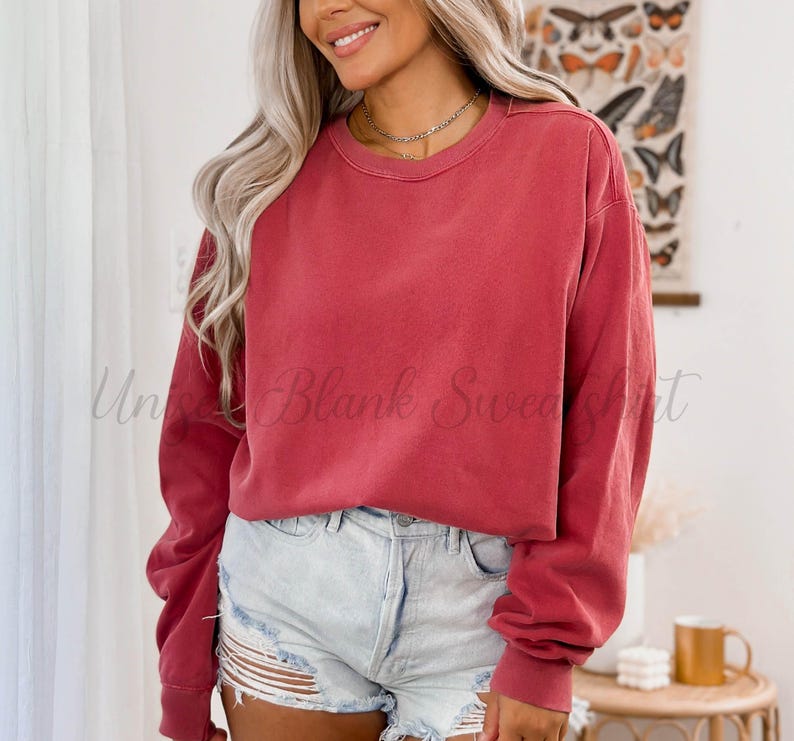 May include: A solid red-orange crewneck sweatshirt with long sleeves. The text "Un Blank Sweatshirt" is visible on the garment. The model is wearing light blue denim shorts. A gold necklace is visible.