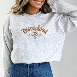 Puede incluir: Sudadera gris claro con la palabra "Disneyland" bordada en marr&oacute;n. Debajo est&aacute; "Est. 1955" y un gr&aacute;fico de Mickey Mouse. La sudadera tiene mangas largas y cuello redondo.