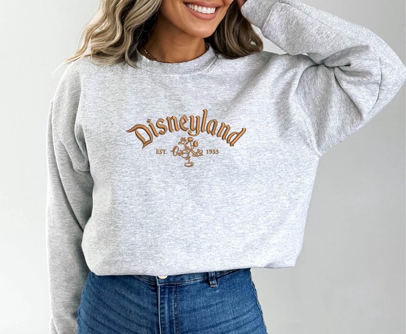 Puede incluir: Sudadera gris claro con la palabra "Disneyland" bordada en marr&oacute;n. Debajo est&aacute; "Est. 1955" y un gr&aacute;fico de Mickey Mouse. La sudadera tiene mangas largas y cuello redondo.