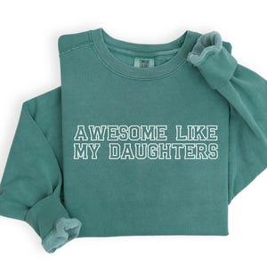 以下が含まれることがあります： 「AWESOME LIKE MY DAUGHTERS」の白い文字が入った、ティール色の長袖スウェットシャツ。スウェットシャツはクルーネックとリブ編みの袖口が付いています。柔らかく快適な素材で作られています。