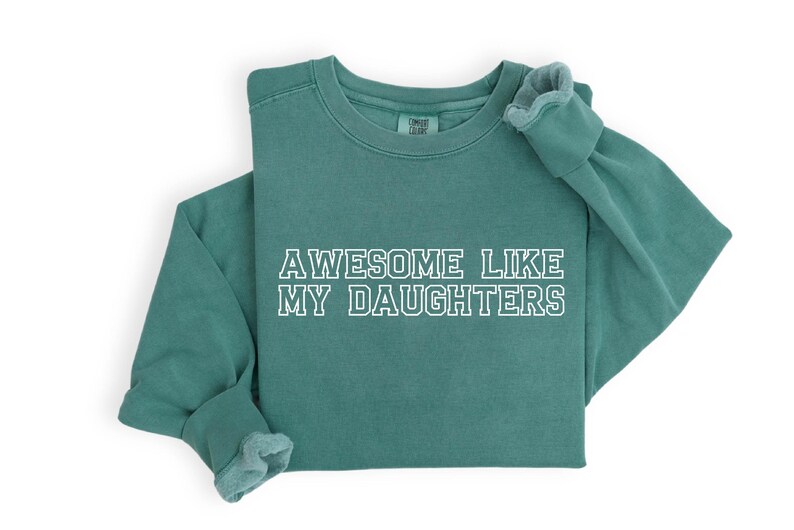 以下が含まれることがあります： 「AWESOME LIKE MY DAUGHTERS」の白い文字が入った、ティール色の長袖スウェットシャツ。スウェットシャツはクルーネックとリブ編みの袖口が付いています。柔らかく快適な素材で作られています。