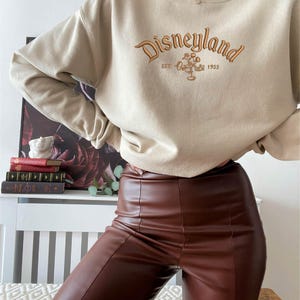 Puede incluir: Sudadera beige con la palabra "Disneyland" bordada en marr&oacute;n, junto con Mickey Mouse y el a&ntilde;o "1955". La sudadera se combina con pantalones marrones de efecto cuero.