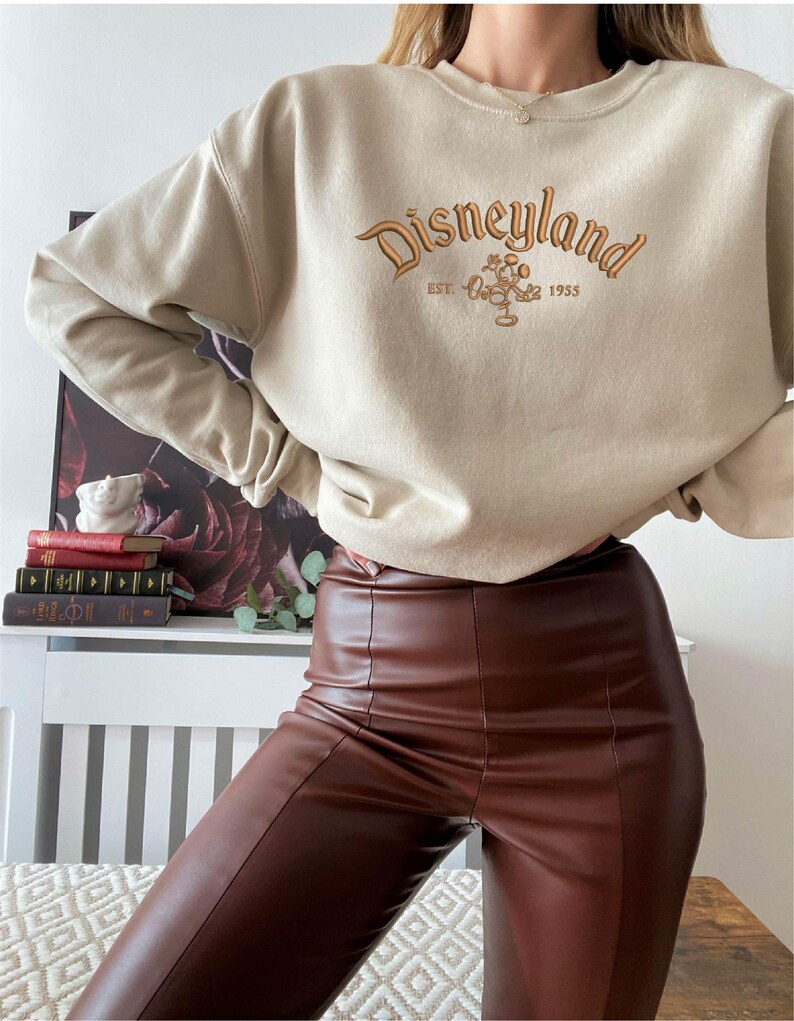 Puede incluir: Sudadera beige con la palabra "Disneyland" bordada en marr&oacute;n, junto con Mickey Mouse y el a&ntilde;o "1955". La sudadera se combina con pantalones marrones de efecto cuero.