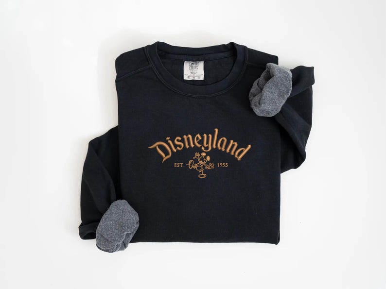 Puede incluir: Sudadera negra con la palabra "Disneyland" bordada en dorado, un dibujo de Mickey Mouse y el a&ntilde;o "1955". Las mangas tienen pu&ntilde;os de tela gris.