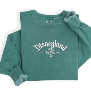 Puede incluir: Sudadera de manga larga de color verde azulado con la palabra "Disneyland" en escritura blanca en el pecho. Debajo, un dibujo de Mickey Mouse y el texto "EST. 1955". La sudadera est&aacute; doblada sobre un fondo blanco.