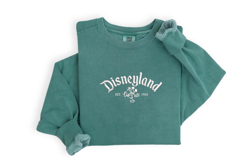 Puede incluir: Sudadera de manga larga de color verde azulado con la palabra "Disneyland" en escritura blanca en el pecho. Debajo, un dibujo de Mickey Mouse y el texto "EST. 1955". La sudadera est&aacute; doblada sobre un fondo blanco.