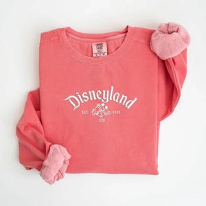 Puede incluir: Sudadera de color coral con la palabra "Disneyland" en escritura blanca, un dibujo de Mickey Mouse y el a&ntilde;o "1955". Las mangas est&aacute;n enrolladas, revelando un forro rosa m&aacute;s claro. Una prenda superior informal y c&oacute;moda.