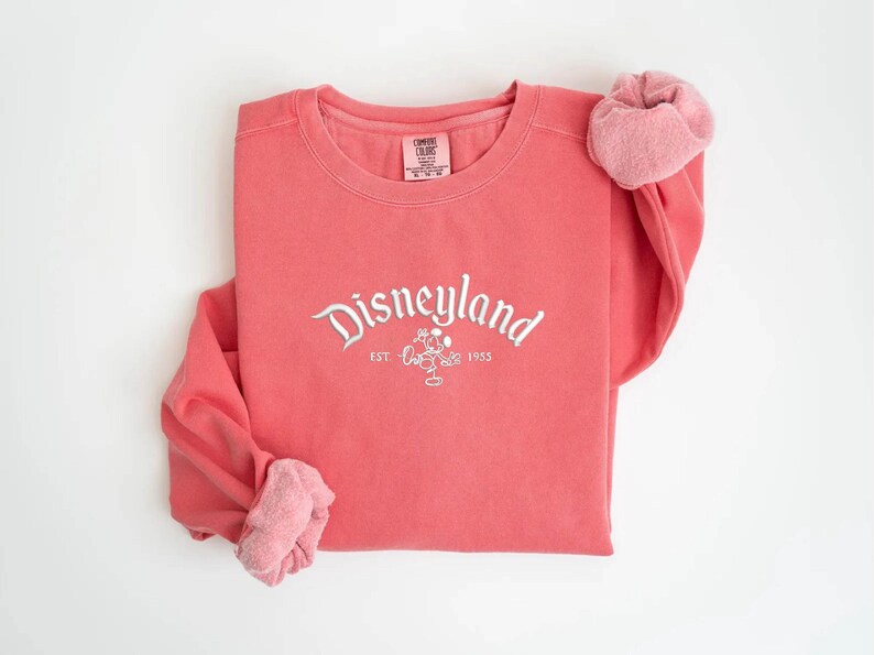 Puede incluir: Sudadera de color coral con la palabra "Disneyland" en escritura blanca, un dibujo de Mickey Mouse y el a&ntilde;o "1955". Las mangas est&aacute;n enrolladas, revelando un forro rosa m&aacute;s claro. Una prenda superior informal y c&oacute;moda.