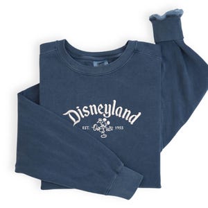 Puede incluir: Una sudadera azul marino de manga larga con la palabra "Disneyland" bordada en escritura blanca en el pecho, junto con un gr&aacute;fico de Mickey Mouse y el a&ntilde;o "1955". La sudadera est&aacute; doblada cuidadosamente sobre un fondo blanco.