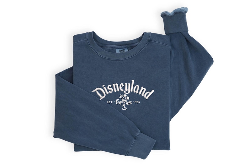Puede incluir: Una sudadera azul marino de manga larga con la palabra "Disneyland" bordada en escritura blanca en el pecho, junto con un gr&aacute;fico de Mickey Mouse y el a&ntilde;o "1955". La sudadera est&aacute; doblada cuidadosamente sobre un fondo blanco.