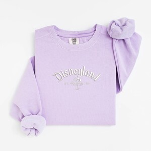Puede incluir: Una sudadera color lavanda con cuello redondo y la palabra "Disneyland" bordada en plata en el pecho. La sudadera tiene mangas largas y un pu&ntilde;o doblado. Las mangas est&aacute;n dobladas en la mu&ntilde;eca. La sudadera es de un material suave.