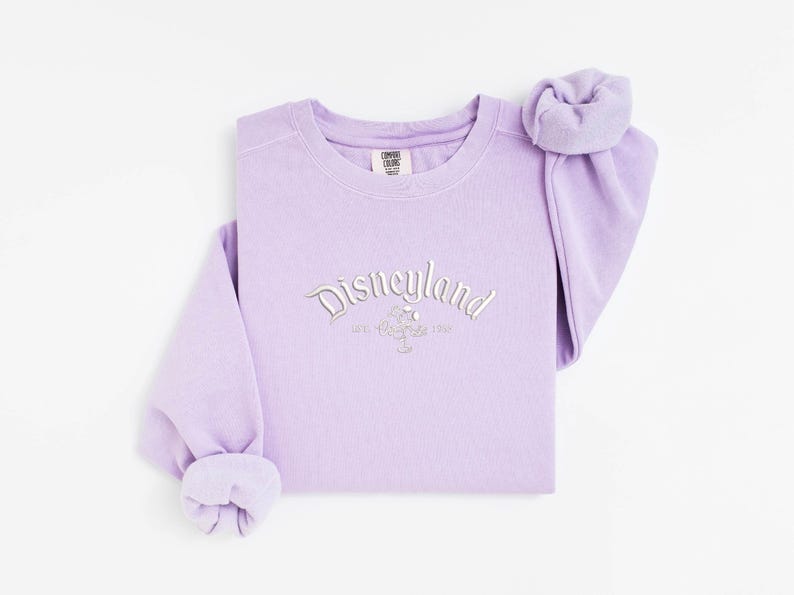 Puede incluir: Una sudadera color lavanda con cuello redondo y la palabra "Disneyland" bordada en plata en el pecho. La sudadera tiene mangas largas y un pu&ntilde;o doblado. Las mangas est&aacute;n dobladas en la mu&ntilde;eca. La sudadera es de un material suave.