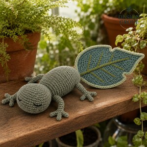 Può includere: Un giocattolo a forma di lucertola lavorato all'uncinetto in verde salvia con un corpo arrotondato e quattro piccole zampe, dotato di una grande coda piatta a forma di foglia in una tonalità verde acqua scuro con venature ricamate verde chiaro.