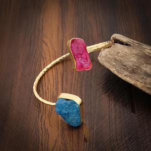 May include: A gold-toned open cuff bracelet featuring a hammered metal band with two raw, irregular-shaped stones at each end. One stone is a vibrant, translucent magenta pink, while the other is a textured, opaque bright blue gemstone.