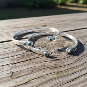Può includere: Due braccialetti a polsino argentati con dettagli in pietra turchese. I braccialetti sono appoggiati su una superficie di legno invecchiato. Il design aperto presenta piccole pietre rotonde. Luce solare.