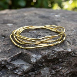 Op de afbeelding: Vier goudkleurige armbanden met een gehamerde textuur liggen op een grijze steen. De armbanden hebben een golvend, organisch ontwerp. De achtergrond is vaag groen. De armbanden zijn van metaal.