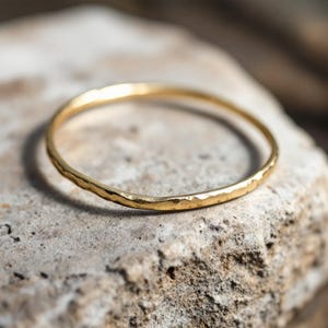 May include: A gold hammered ring, displayed on a textured stone. The ring is a simple band with a slightly uneven surface, catching the light. The ring's design is minimalist and elegant, suitable for everyday wear.