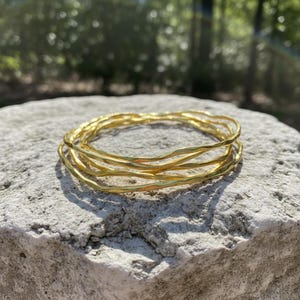 Op de afbeelding: Een stapel van vijf goudkleurige armbanden met een getextureerde, gehamerde uitstraling. De armbanden liggen op een lichtgrijze stenen ondergrond, met een vage groene achtergrond van bomen en zonlicht.