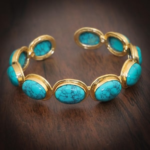Könnte beinhalten: Goldfarbenes Armreif-Armband mit ovalen Türkissteinen. Das Armband hat eine Reihe von Türkissteinen, die in goldfarbenen Fassungen gefasst sind. Die Steine sind leuchtend türkis mit dunklen Adern. Das Armband wird auf einer dunklen Holzoberfläche präsentiert.