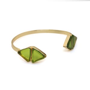 May include: A gold-toned open cuff bracelet featuring two triangular, translucent lime green glass accents at each end. The metal band is smooth and polished, connecting to the geometric green glass pieces that face inward.