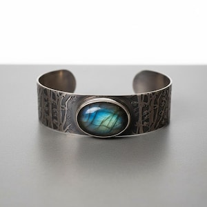 May include: A silver cuff bracelet with an oval, iridescent blue and green gemstone. The bracelet band features a detailed forest design. The gemstone is set in a silver bezel, creating a striking contrast.