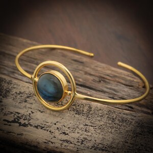 May include: Gold-toned cuff bracelet featuring a round, iridescent blue-green stone set within a circular design. The bracelet has a simple, open design with a polished finish, suitable for everyday wear.