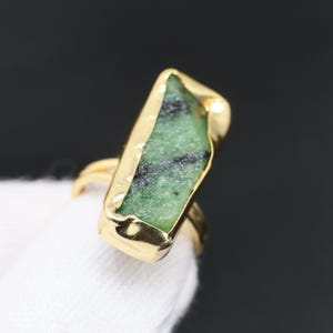 Puede incluir: Un anillo de tono dorado con una piedra verde de corte irregular y forma rectangular con inclusiones oscuras, engastada en un bisel de metal liso y pulido. La banda es delgada y metálica, resaltando la textura natural de la piedra.