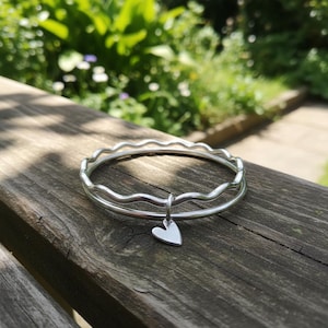 Puede incluir: Una pulsera de plata con un diseño ondulado y un pequeño dije de corazón. La pulsera está sobre una superficie de madera desgastada, con un fondo verde borroso.