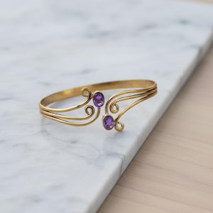 May include: A gold-colored metal bracelet with two oval, purple gemstones. The bracelet has a delicate, open design with swirling accents. The gemstones are set in the center of the bracelet.