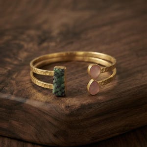 May include: A gold-toned open cuff bracelet featuring a hammered metal texture. One end of the cuff is adorned with a rectangular green and black speckled stone, while the other end features two small, light pink teardrop-shaped stones.
