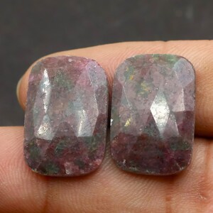 May include: Two faceted, rectangular gemstones with a mix of pink, purple, and green hues. The gems are held in a hand, showcasing their polished surfaces and internal patterns. The stones are likely for jewelry making.