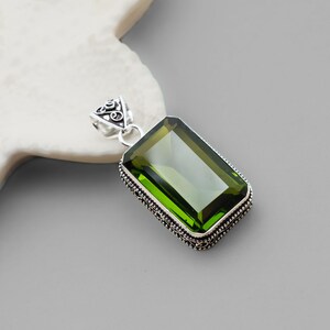 May include: A rectangular pendant with a large, vibrant green gemstone. The gemstone is set in a silver-colored metal frame with intricate detailing. The pendant has a decorative bail with a loop for a chain.
