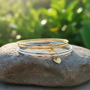 Op de afbeelding: Drie armbanden, twee zilveren en één gouden, liggen op een grijze steen. De gouden armband heeft een gehamerde textuur. Elke armband heeft een kleine gouden hartbedel. De achtergrond is zachtgroen met zonlicht.