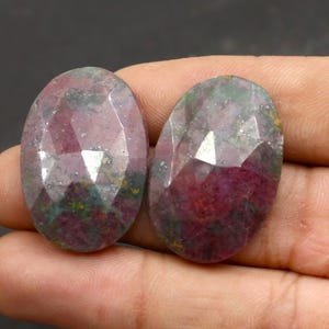 Op de afbeelding: Twee ovale, gefacetteerde edelstenen. De edelstenen hebben een dieprode kleur met groene en grijze vlekken. De facetten creëren een sprankelend effect. De stenen worden in een hand tegen een donkere achtergrond gehouden. Geschikt voor het maken van sieraden.
