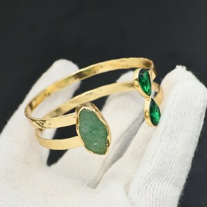 May include: A gold-toned open cuff bracelet featuring a hammered metal finish. One end of the cuff is adorned with a large, irregular, matte light green stone, while the other end features two smaller, faceted, teardrop-shaped emerald green stones.