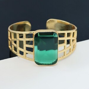 May include: A gold-toned open cuff bracelet featuring a large, rectangular, emerald-green faceted gemstone at the center. The band is constructed with a geometric grid pattern, creating an openwork design that wraps around the wrist.