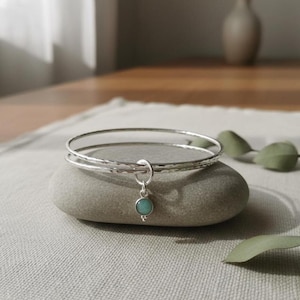May include: Two silver bangles with a hammered texture rest on a gray stone. A small charm with a turquoise-colored stone hangs from the bangles. The jewelry is displayed on a neutral-colored surface.
