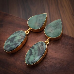 May include: A pair of drop earrings featuring two green and black mottled stones set in gold-toned metal. Each earring consists of a teardrop-shaped stone at the top connected by a small gold ring to a larger oval-shaped stone at the bottom.