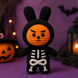 May include: A crochet plush toy featuring a round orange face with an angry expression, wearing a black bunny-eared hood and a black skeleton-patterned body suit with white stitched rib cage and pelvic bone details on the front.