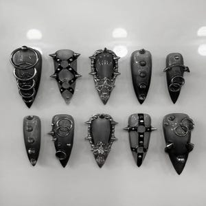 Peut inclure: Collection de dix modèles de nail art gris de style gothique. Chaque ongle présente des ornements uniques, notamment des anneaux métalliques, des clous et des filigranes ornés. Les ongles sont pointus et ont un aspect sombre et fumé.