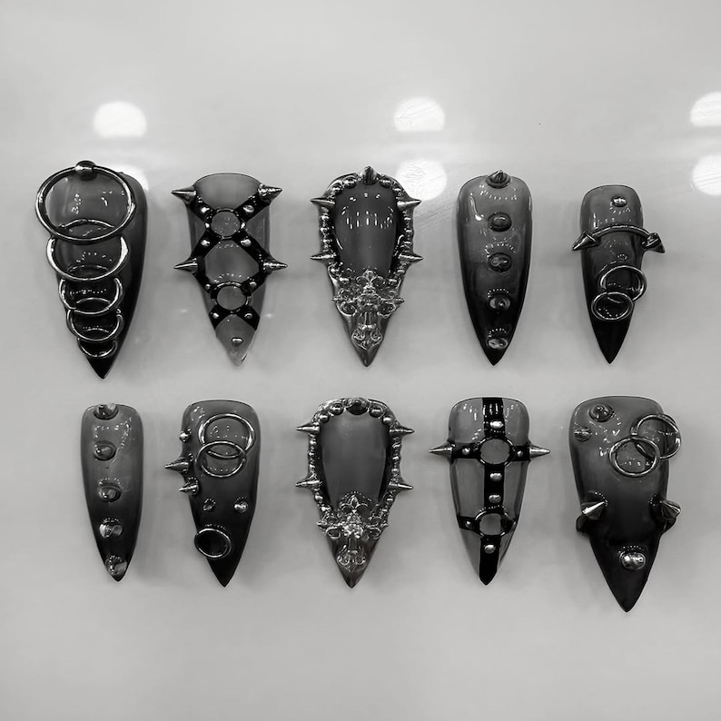 May include: Ten grey, gothic-style nail art designs are displayed. Each nail features unique embellishments, including metal rings, studs, and ornate filigree. The nails are pointed and have a dark, smoky appearance.