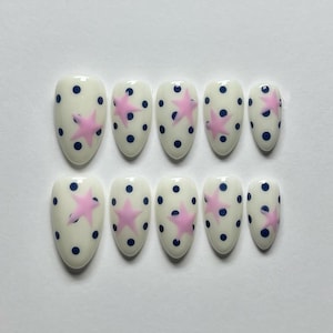 May include: Set of ten oval-shaped false nails. Each nail is off-white with a pattern of navy blue polka dots and a pink star. The nails are arranged in two rows of five.