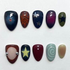 May include: A set of ten artificial nails with various designs. The nails feature colors like navy, burgundy, olive green, and nude. Designs include stars, swirls, and geometric shapes. Some nails have silver accents.
